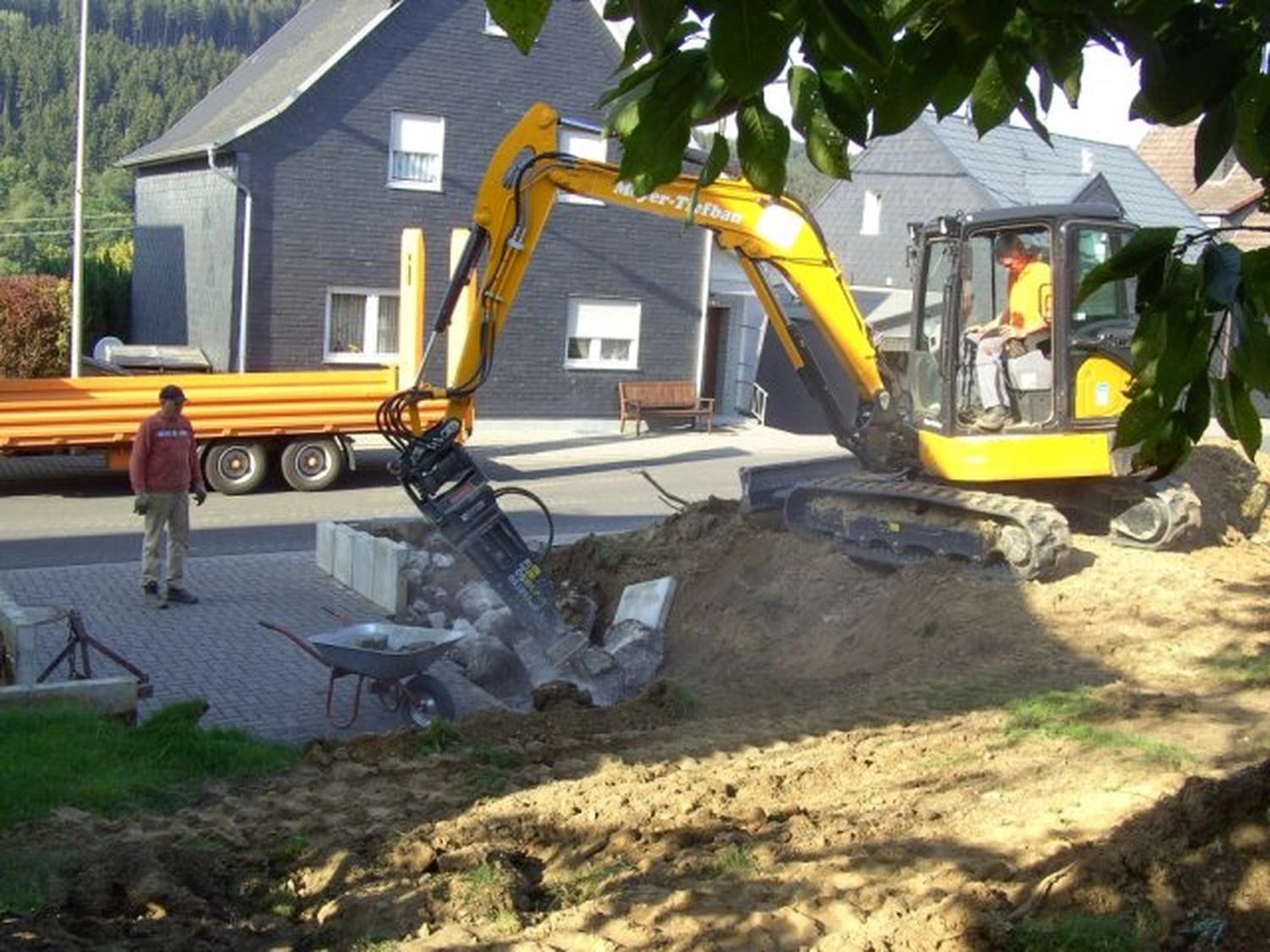 Hans Werner Meyer GmbH - Tiefbauunternehmen - Ein gelber Minibagger mit hydraulischem Abbruchhammer bricht Beton in einem Wohngarten auf, während ein Arbeiter daneben steht.