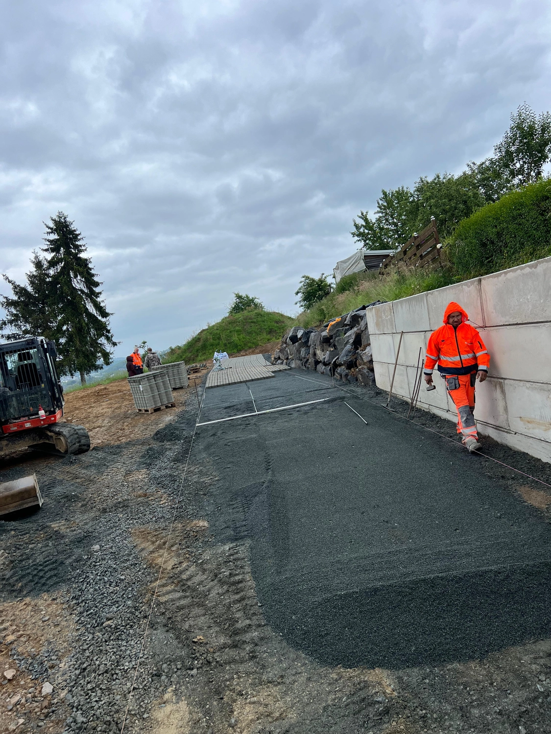 Hans Werner Meyer GmbH - Tiefbauunternehmen - Unter bewölktem Himmel bereiten Bauarbeiter eine Straße für die Pflasterung vor, indem sie eine Schicht aus Kies und gestapelten Pflastersteinen auftragen.
