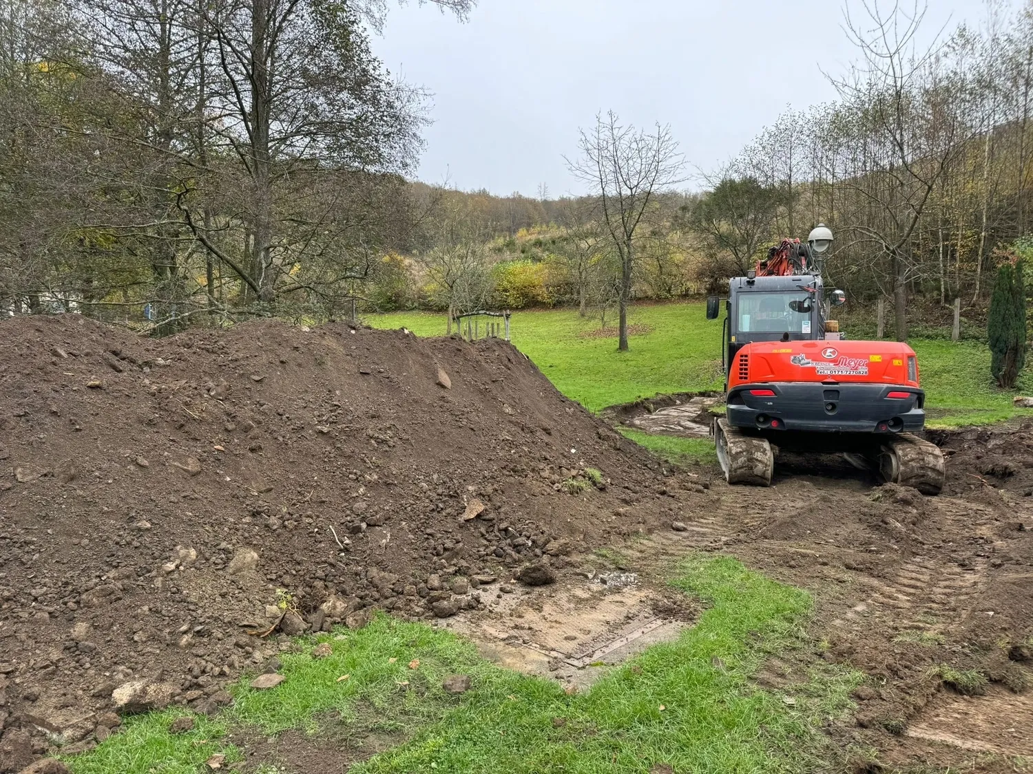 Hans Werner Meyer GmbH - Tiefbauunternehmen - Ein orangefarbener Meyer-Bagger steht auf einem Feldweg in einem grasbewachsenen Feld neben einem großen Erdhügel und mehreren kahlen Bäumen.