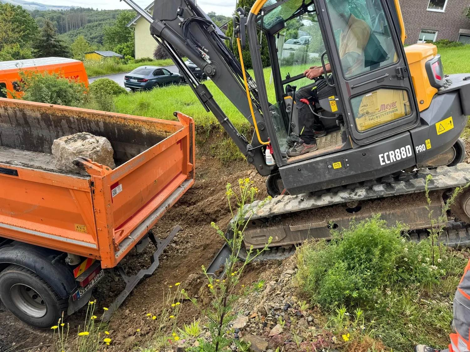 Hans Werner Meyer GmbH - Tiefbauunternehmen - Ein Volvo ECR88D Kompaktbagger verlädt schwere Steine auf die Ladefläche eines orangefarbenen Muldenkippers auf einer Wohnbaustelle.
