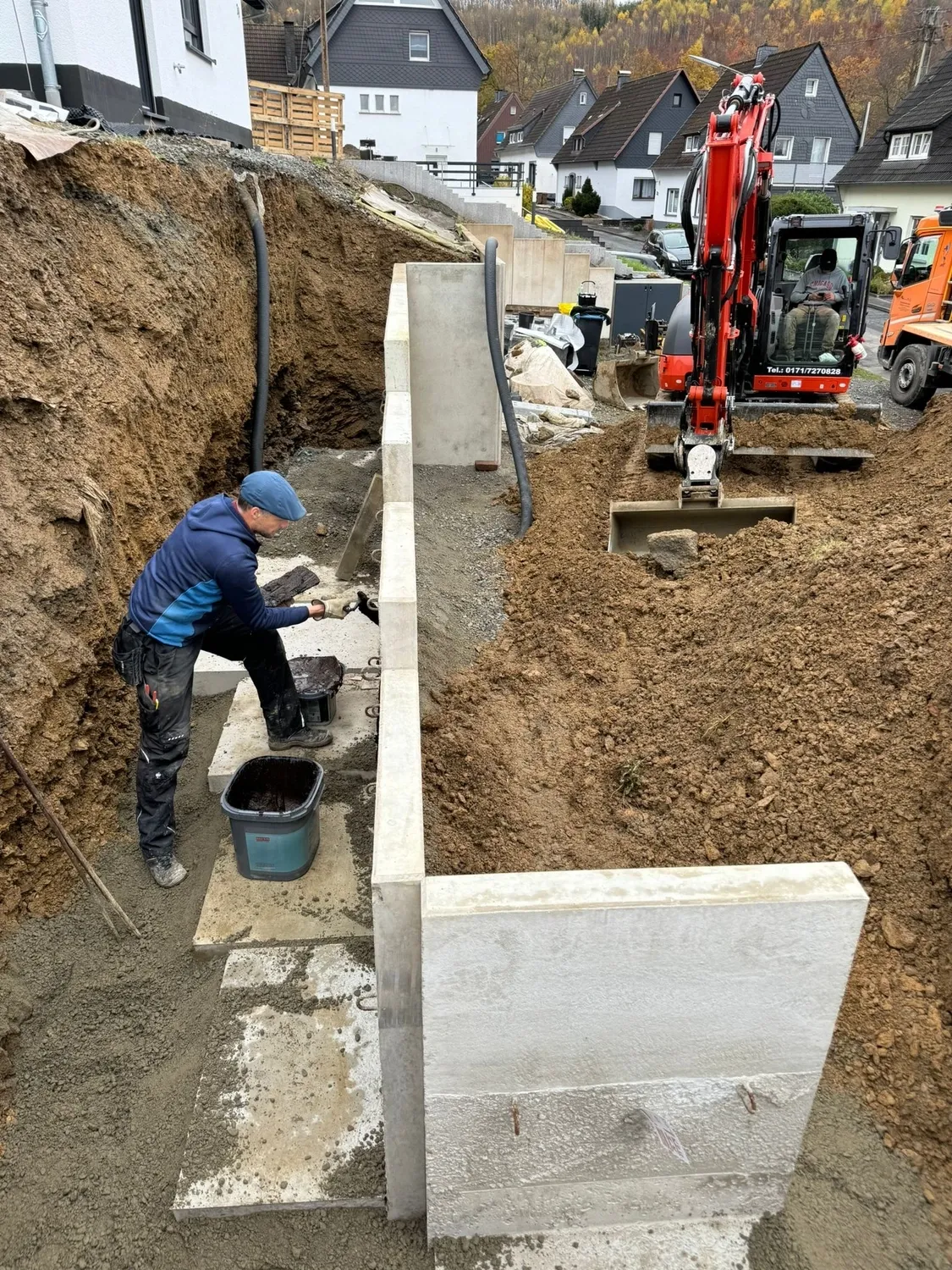 Hans Werner Meyer GmbH - Tiefbauunternehmen - Bau einer Betonstützmauer an einem Hang mit einem Bagger und einem Arbeiter, der Dichtmittel aufträgt.