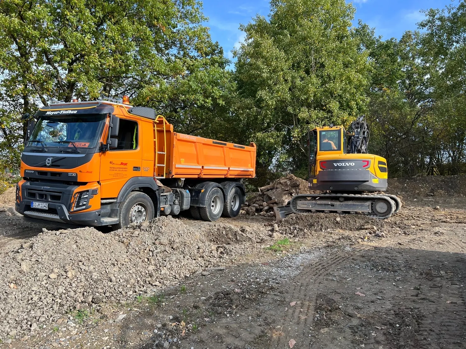 Hans Werner Meyer GmbH - Tiefbauunternehmen - Ein orangefarbener Volvo-Muldenkipper wird auf einer bewaldeten Baustelle von einem gelb-grauen Volvo-Kompaktbagger mit Erde beladen.